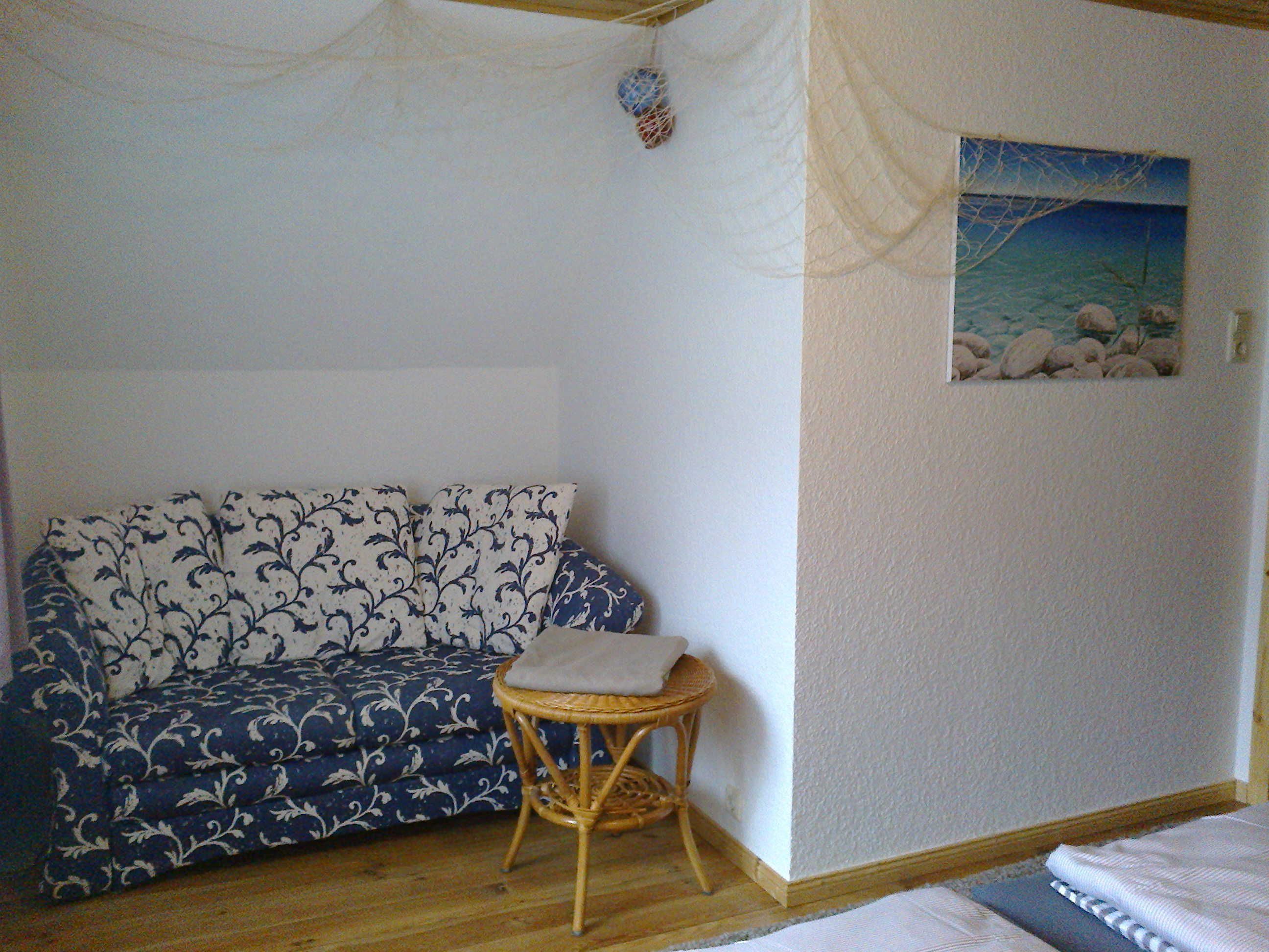 A room with a patterned sofa, a small wicker chair, a painting of a beach scene, and a bed with a striped bedspread.
