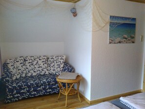 A room with a patterned sofa, a small wicker chair, a painting of a beach scene, and a bed with a striped bedspread.