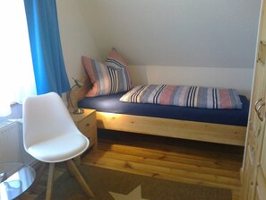 A small, single-bed room with a wooden floor, a white chair, a small table, and a window with blue curtains.
