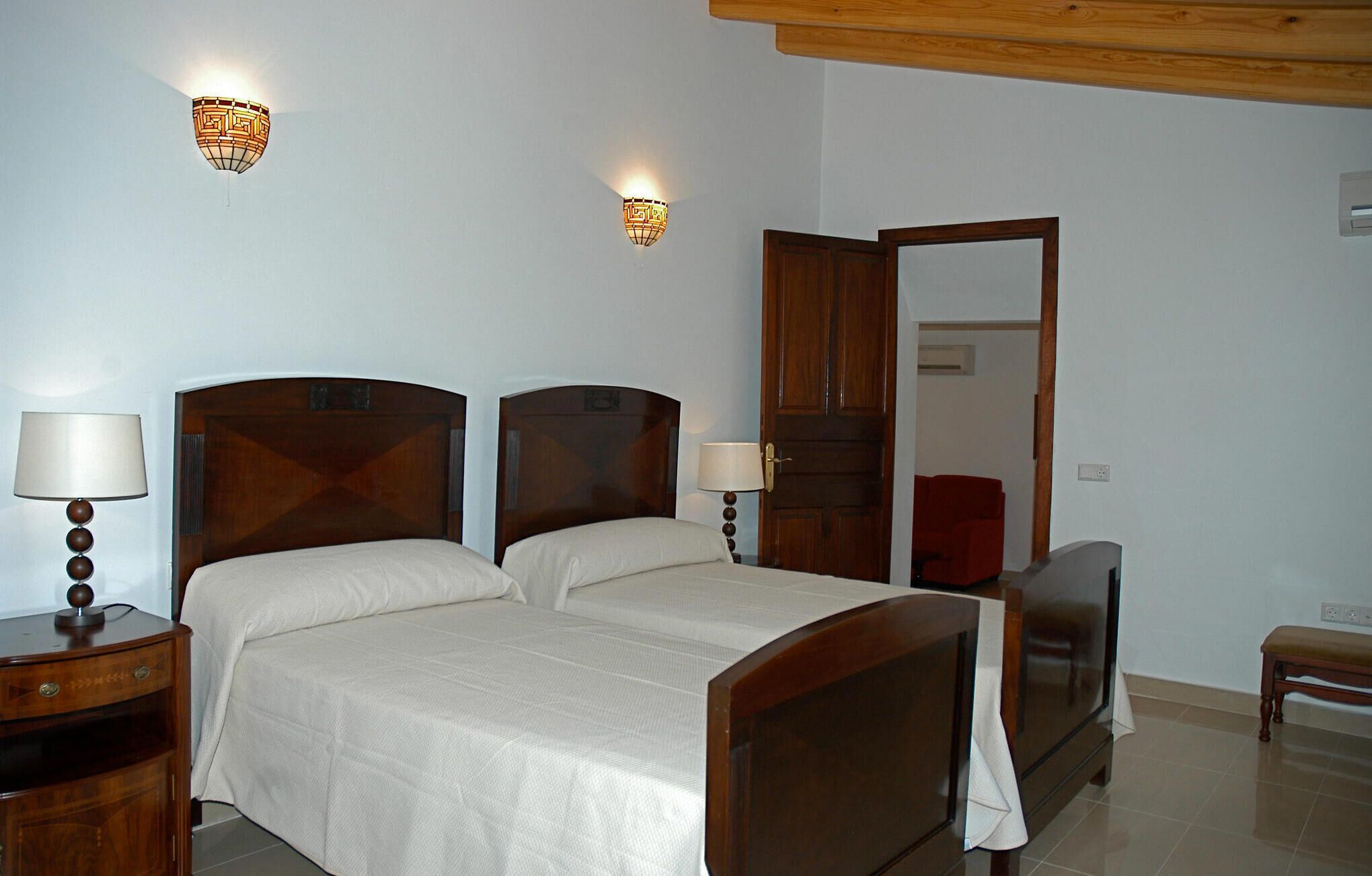 A double bed with wooden headboards, two bedside lamps, a wooden nightstand, and a red chair in the background.