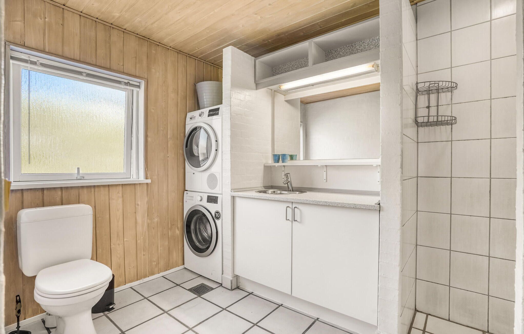 Une salle de bain moderne et compacte comprenant des toilettes, un lave-linge et un sèche-linge superposés, un petit lavabo et un espace douche.