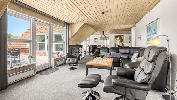 A modern living room with a large sectional sofa, a coffee table, and a dining area with a table and chairs. There is a large window providing a view of the outside. - Cozy home in Blåvand with sauna (Blåvand)