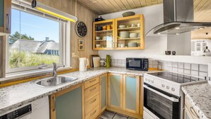 A modern kitchen with wooden cabinets, a built-in oven, a microwave, and a large window offering a view of the outdoors. - Cozy home in Blåvand with sauna (Blåvand)