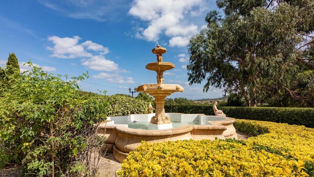 A multi-tiered stone fountain surrounded by lush greenery and yellow flowers. - Awesome home in Sineu (Sineu)