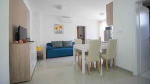A modern living room with a blue sofa, a dining table with chairs, a TV, and a kitchen area with cabinets and appliances. - Holiday flat with shared pool and barbecue (Baška)