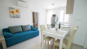A modern living room with a blue sofa, a dining table with chairs, and a kitchen area with a refrigerator and stove. - Holiday flat with shared pool and barbecue (Baška)