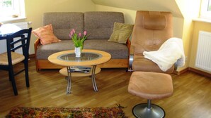 A cozy living room with a sofa, a chair, a small table with a vase of flowers, and a window. - Apartment Zum Kölpinsee - K��lp - Will (Loddin)