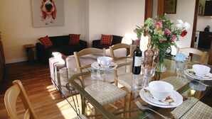 A dining area with a glass table set for four, surrounded by chairs. There is a sofa with cushions in the background, and a painting on the wall.