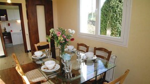 A dining area with a glass table set for four, surrounded by wooden chairs with patterned cushions. A vase with flowers is on the table, and a window provides a view of greenery outside.