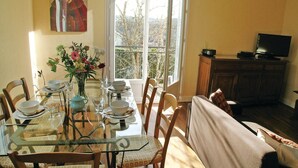 A dining area with a glass table, chairs, and a vase of flowers. There is a window with a view of trees and a television on a cabinet.