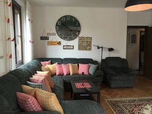 Ein Wohnzimmer mit einer großen, runden Wanduhr, einem Sofa mit vielen Kissen, einem kleinen Tisch mit Fernbedienung und einer Stehlampe.