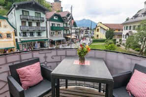 A balcony with wicker furniture and a table, overlooking a town square with buildings and a mountain in the background.