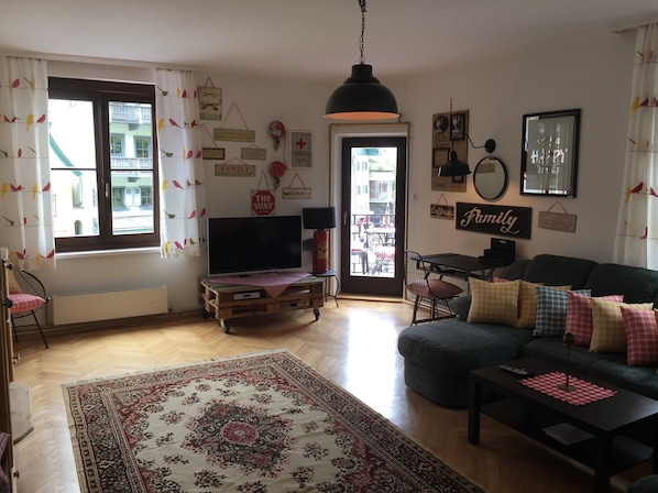 Ein Wohnzimmer mit Fernseher, Sofa und Couchtisch. Eine Tür führt nach draußen, und der Raum ist mit einem Teppich und Wandbildern dekoriert.