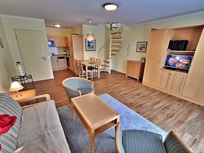 Un espacio habitable compacto con comedor, sofá, televisor y una escalera que lleva a una cocina.