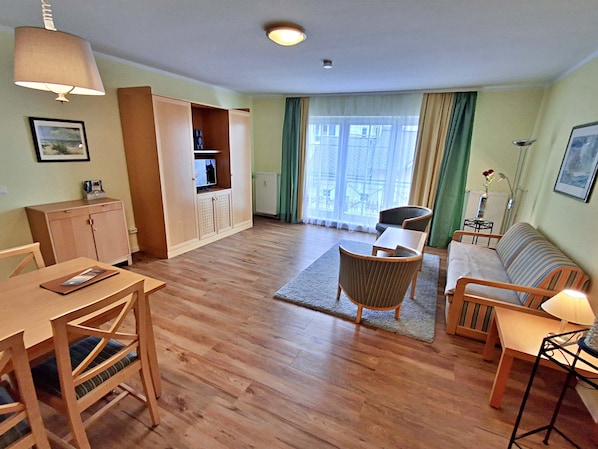 A living room with a wooden floor, a sofa, a dining table with chairs, a TV on a cabinet, and a window with curtains.
