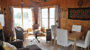 Una habitación acogedora con paredes de madera, una mesa redonda y cuatro sillas. Hay dos ventanas con cortinas, una lámpara y un colcha decorando la pared.