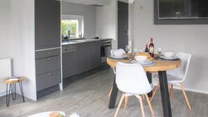 Una cocina moderna con armarios grises, una mesa de comedor de madera y sillas blancas. Hay una ventana que proporciona luz natural y un televisor de pantalla plana montado en la pared.
