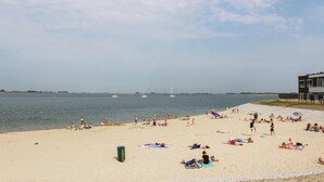 Una playa con gente tomando el sol y nadando, un muelle con un edificio, y veleros en el agua.