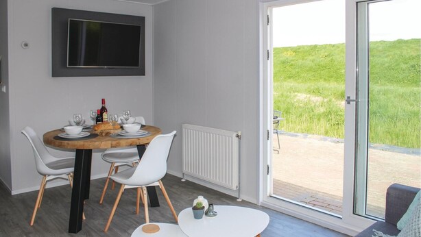 Un comedor con una mesa redonda, sillas blancas, una mesita auxiliar, un televisor montado en la pared y una puerta corrediza de vidrio que da a un área exterior.