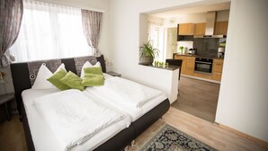 A modern hotel room with a large bed, a small desk, and a view into the kitchen area.