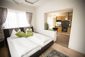 A modern hotel room with a large bed, a small desk, and a view into the kitchen area.