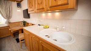 Una cocina con armarios de madera, un lavamanos, una ventana con cortinas y una zona de comedor con una silla y mesa.
