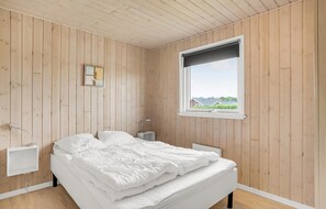 A bedroom with wooden walls, a bed, a bedside table, a lamp, and a window with a view of houses. - Gorgeous home in Tarm with sauna (Tarm)