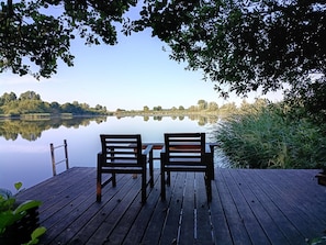 Une terrasse en bois avec deux chaises donnant sur un lac paisible, entourée d’arbres et de végétation.