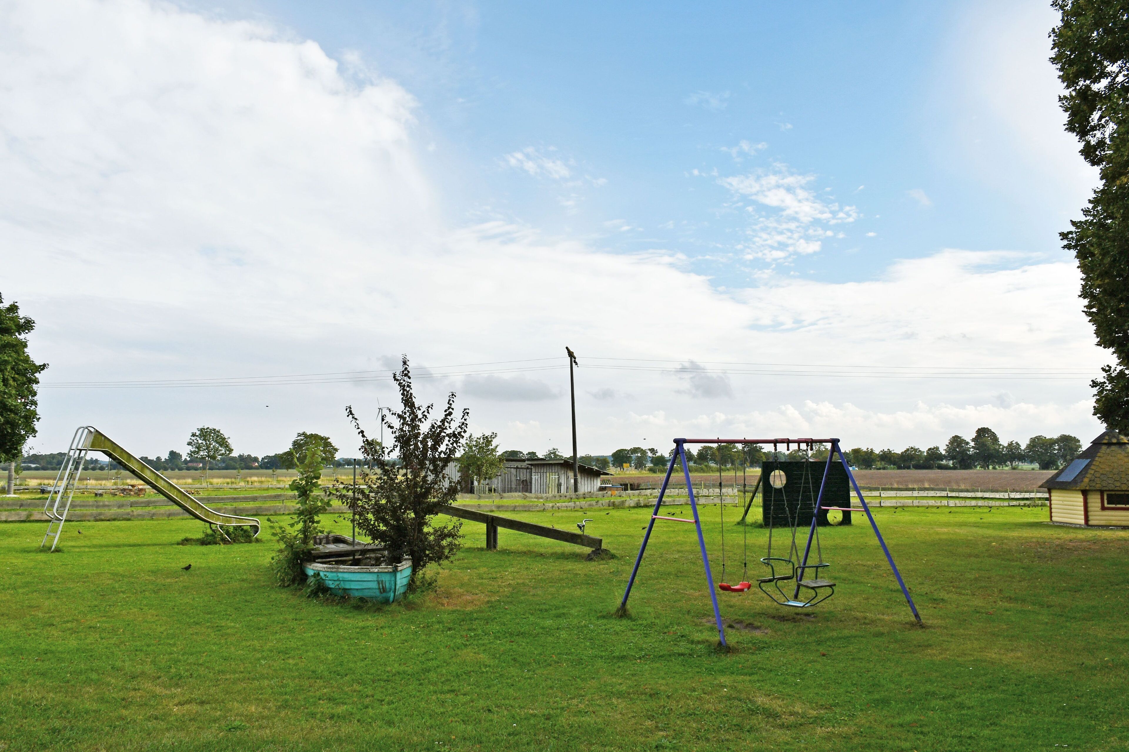 Un parque infantil con columpios, un tobogán y un balancín en un campo cubierto de hierba, con un granero y árboles al fondo.