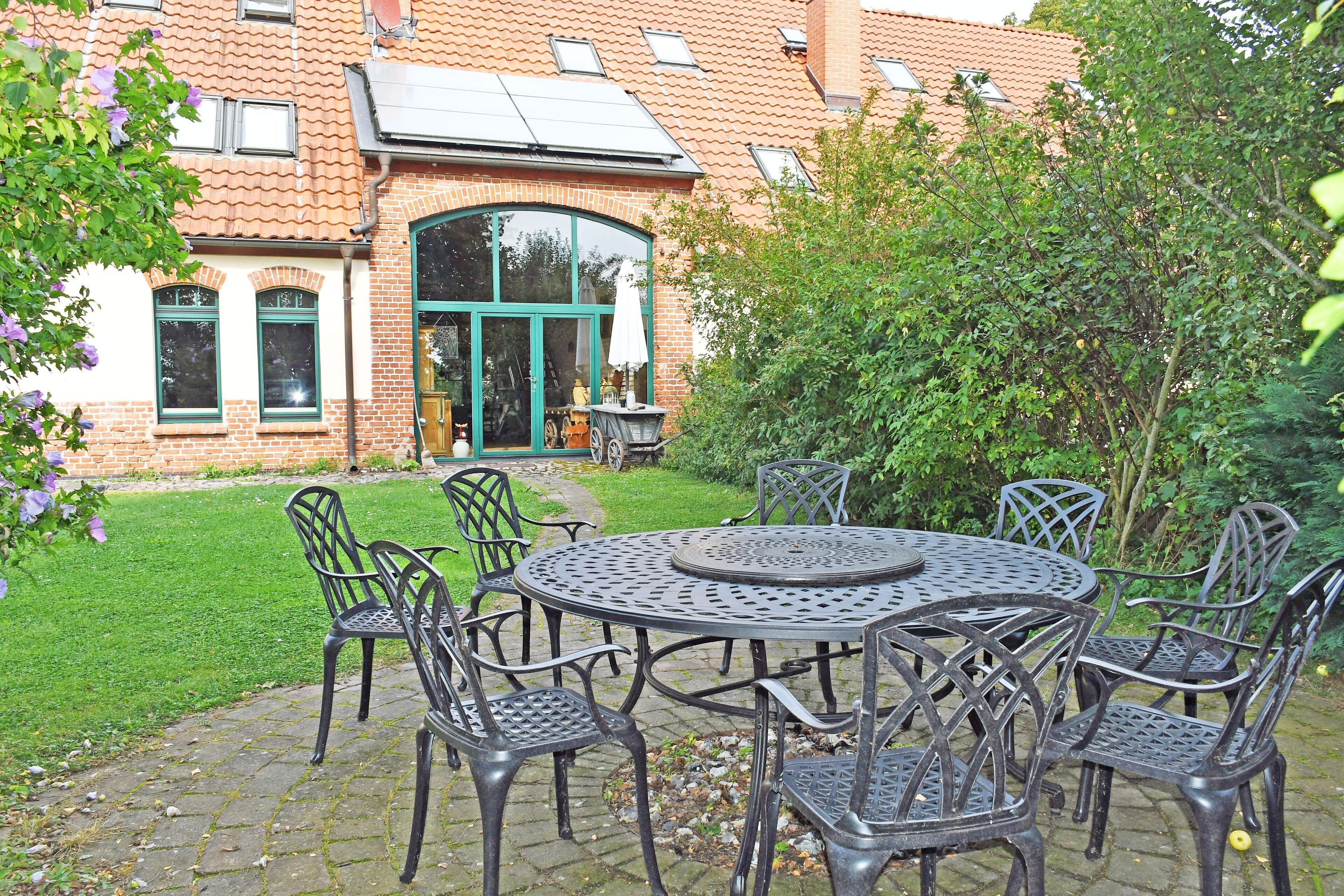 Een terras met een ronde metalen tafel en stoelen, een bakstenen gebouw met een groene deur op de achtergrond, en een verzorgd gazon.