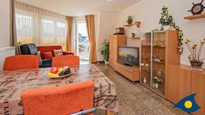 Ein Wohnzimmer mit einer Couch, einem Esstisch mit Obstschale, einem Fernseher und einer Kommode mit Dekoration.