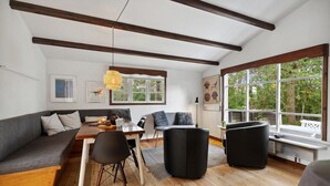 Ett modernt vardagsrum med trägolv, en grå soffa, ett matbord med stolar, en svart pall och stora fönster med utsikt över grönområden.