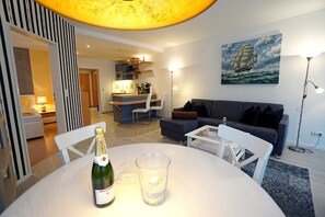 A modern living room with a dining table, a bottle of champagne, and a glass. There is a sofa, a lamp, and a painting on the wall.