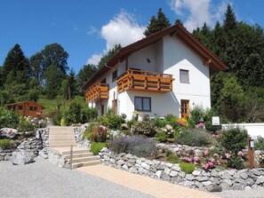 A two-story chalet with a wooden balcony, surrounded by a landscaped garden with various plants and flowers. - Apartment am Rosengarten - Rosengarten (Füssen)