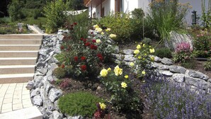 A garden with a variety of flowers and a stone pathway leading to a building with a wooden balcony. - Apartment am Rosengarten - Rosengarten (Füssen)