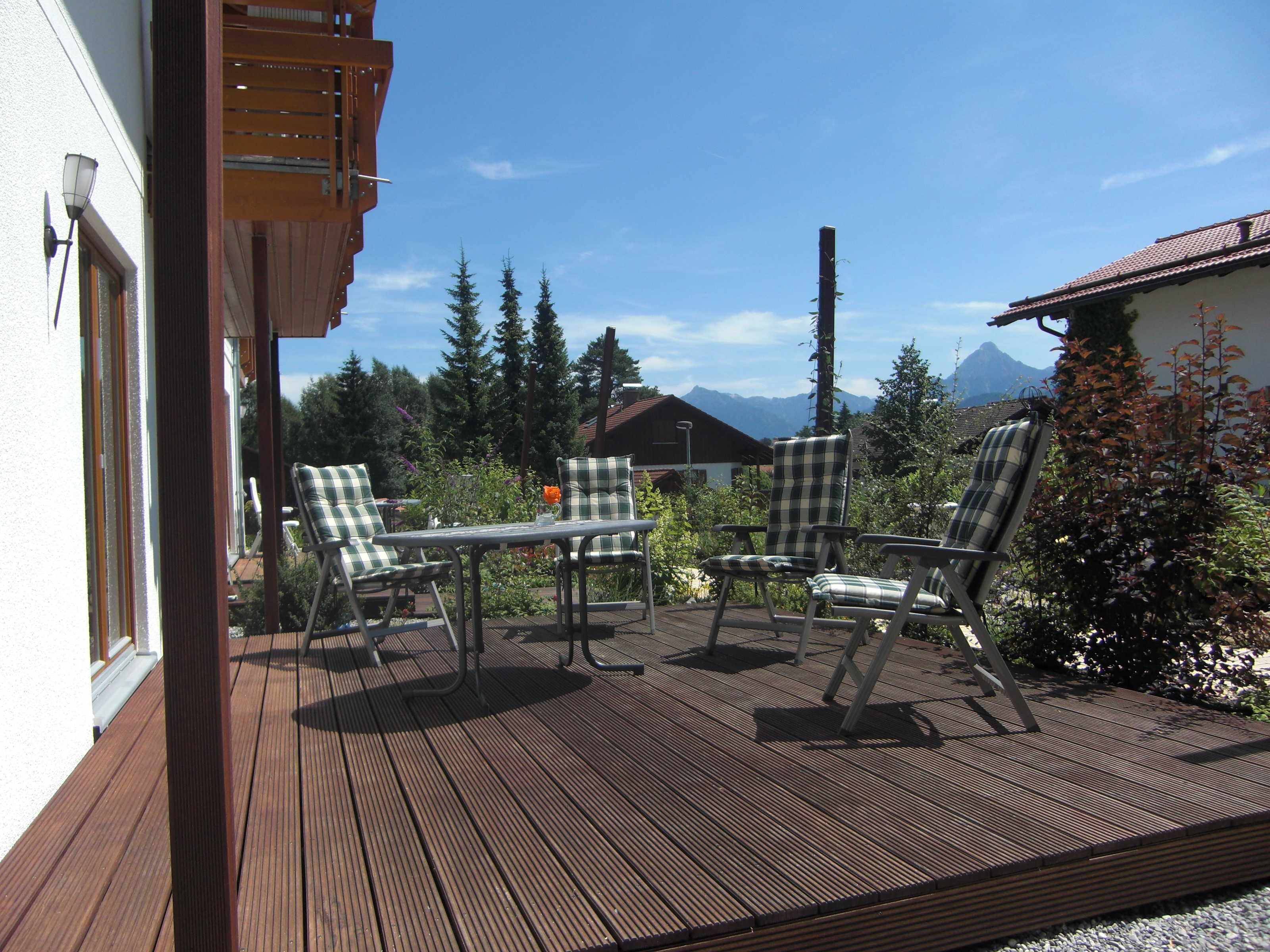 En udendørs terrasse med et bord og fire stole, omgivet af grøn vegetation og udsigt til et bjerg.