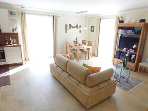 A living room with a sofa, a dining area with chairs, a kitchen with a counter, and a TV stand with speakers.