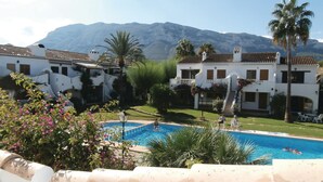 A pool area with a mountain backdrop, surrounded by white buildings with terracotta roofs and palm trees.
