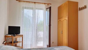 Un dormitorio con cama, televisor sobre un mueble de madera, armario y ventana con cortinas traslúcidas.