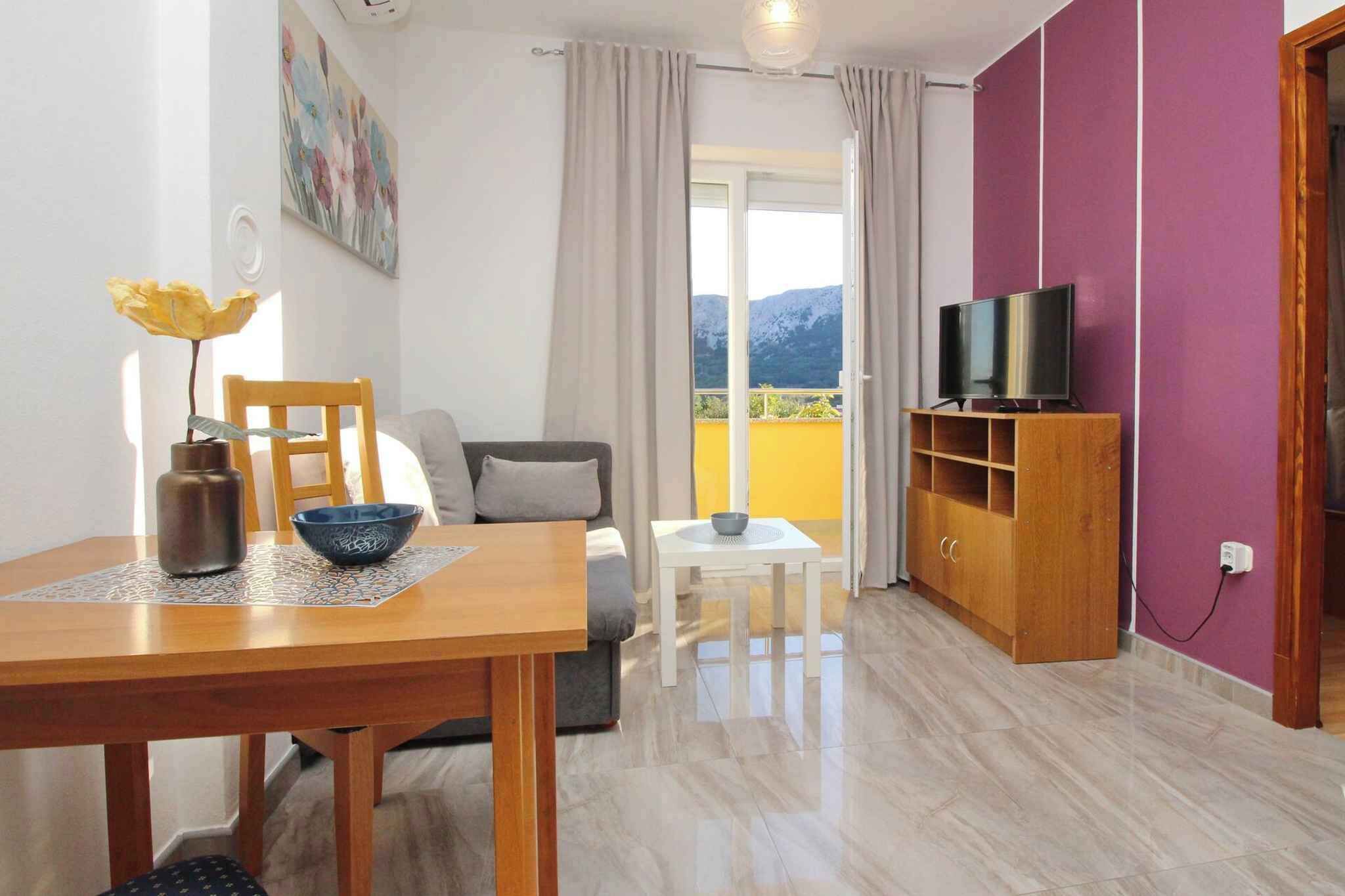 A living room with a sofa, a dining table with a vase and bowl, a TV on a wooden stand, and a balcony with a mountain view.