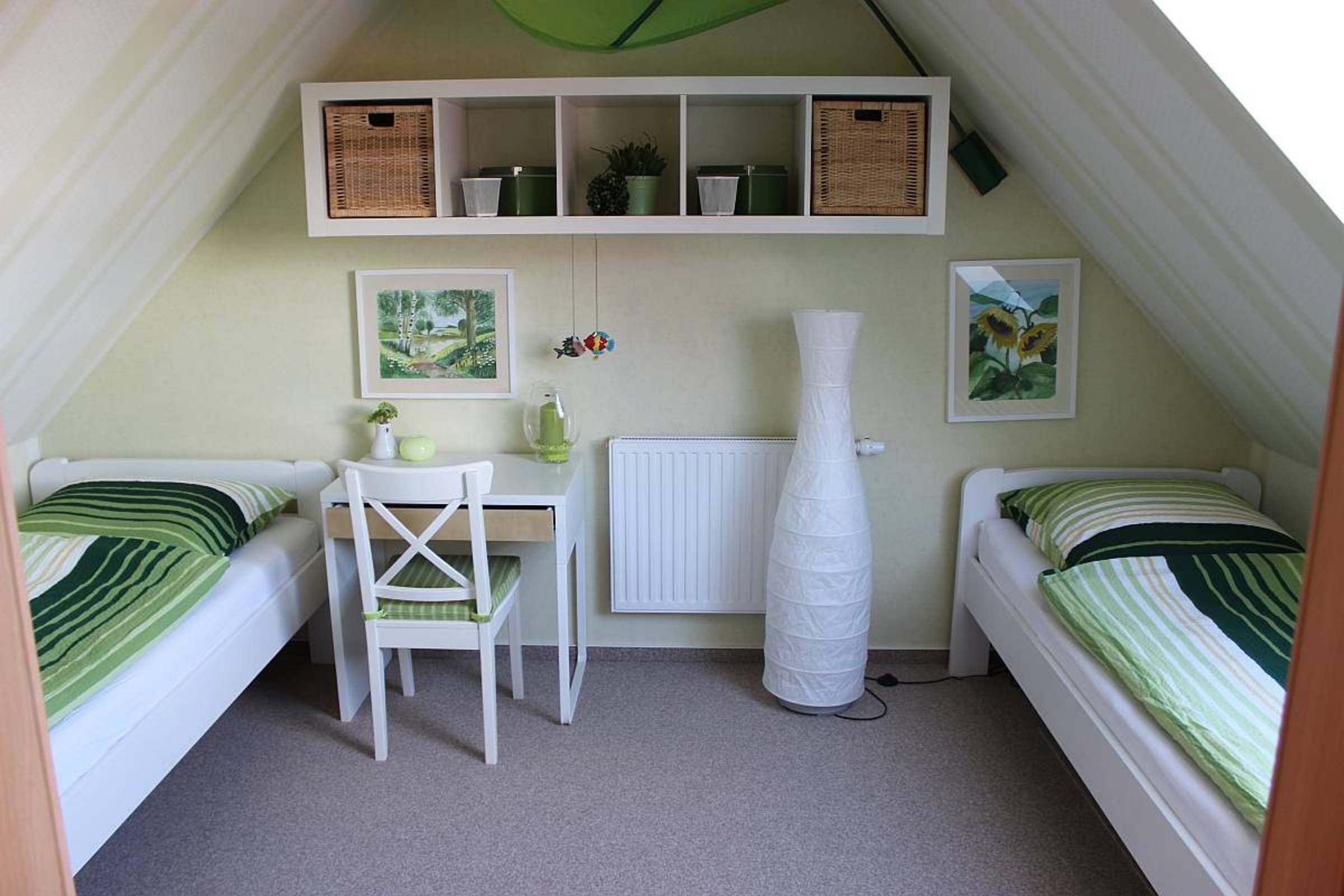 Ein kleines, dreieckiges Schlafzimmer im Dachgeschoss mit zwei Einzelbetten, einem Schreibtisch und einem Stuhl. Über den Betten befinden sich Regale mit Körben und einer Topfpflanze.