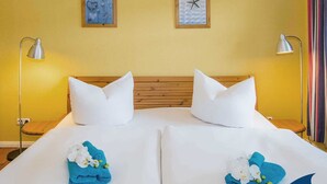 A hotel room with two beds, white linens, and blue towels with flowers. There are wall art pieces above the beds and two bedside lamps.