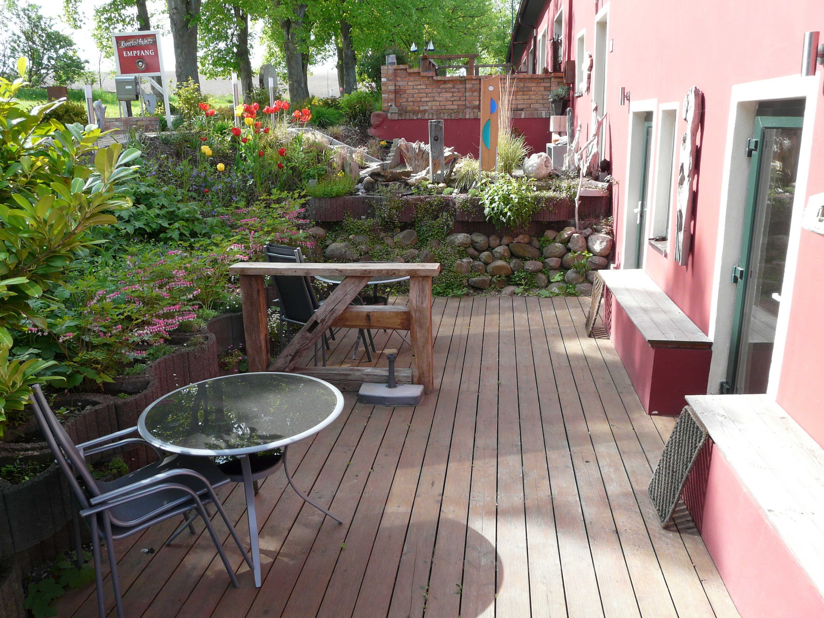 Un terrazzo in legno con tavolo e sedie, circondato da un giardino con diverse piante e fiori.
