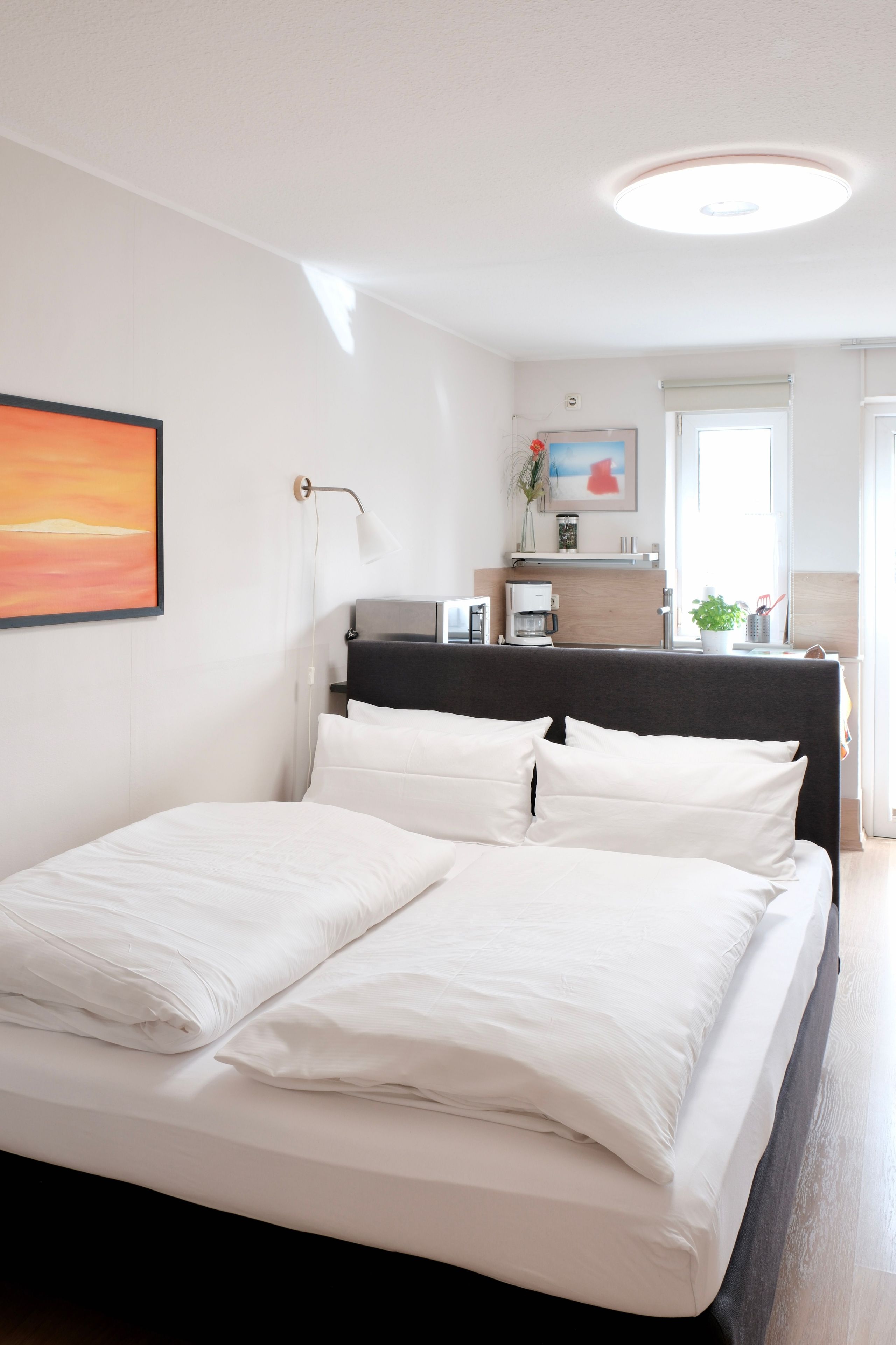 Un lit bien fait, avec du linge de lit blanc, une lampe de chevet, un tableau encadré au mur et une fenêtre avec des stores.