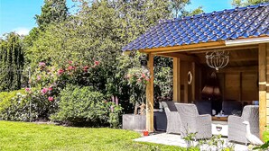 Un'area salotto esterna coperta con mobili in vimini, una pergola con tetto in tegole e un giardino con diverse piante e fiori.