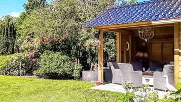 Un'area salotto esterna coperta con mobili in vimini, una pergola con tetto in tegole e un giardino con diverse piante e fiori.