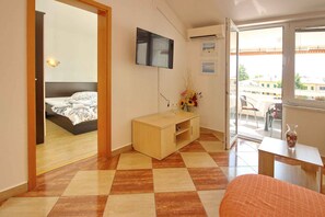 A hotel room with a bed, a wooden desk, a TV mounted on the wall, and a balcony with a view of buildings.