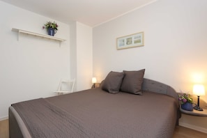 A neatly arranged bedroom with a large bed, a bedside table with a lamp, a small shelf with a plant, and a framed picture on the wall.