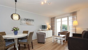 A modern living room with a round dining table, wicker chairs, a sofa, a TV on a cabinet, and large windows with curtains.