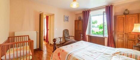 Een slaapkamer met een houten bed, een stoel, een radiator, een raam met gordijnen en een deur die naar een andere kamer leidt.